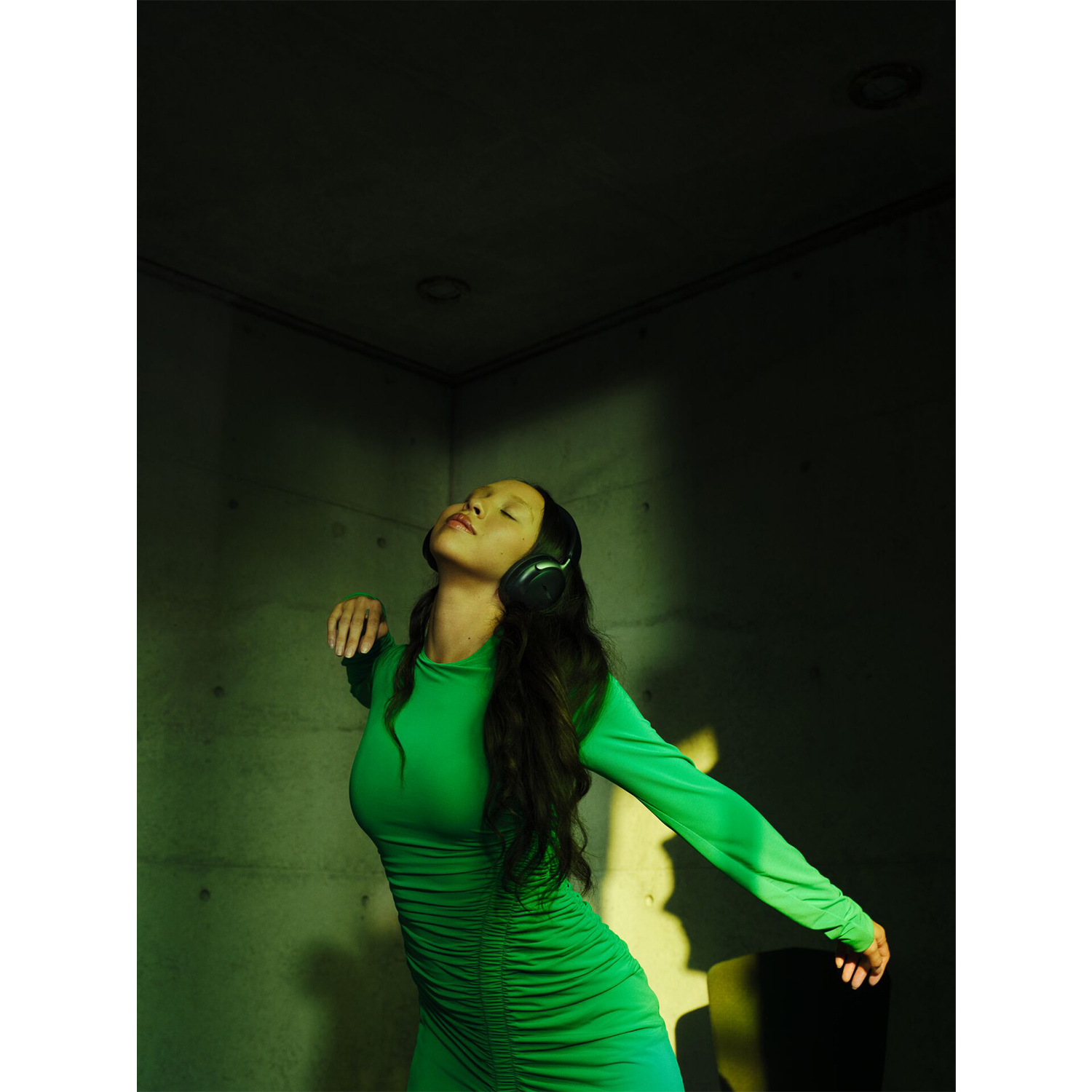 Donna in abito verde con cuffie, occhi chiusi, braccia tese, in una stanza illuminata di verde.