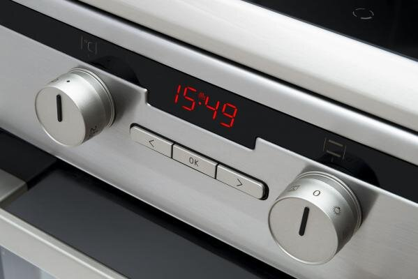 Zbliżenie panelu sterowania piekarnika z cyfrowym zegarem wyświetlającym 15:49, z pokrętłami i przyciskami.