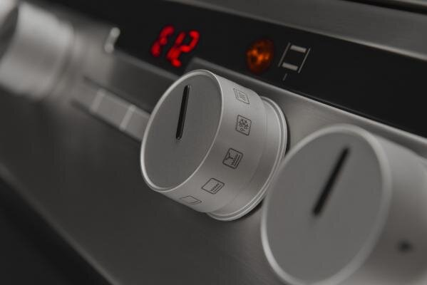 Zbliżenie elementów sterowania piekarnika. Dwa srebrne pokrętła z ikonami. Czerwony wyświetlacz cyfrowy pokazuje '12'. Szare tło.