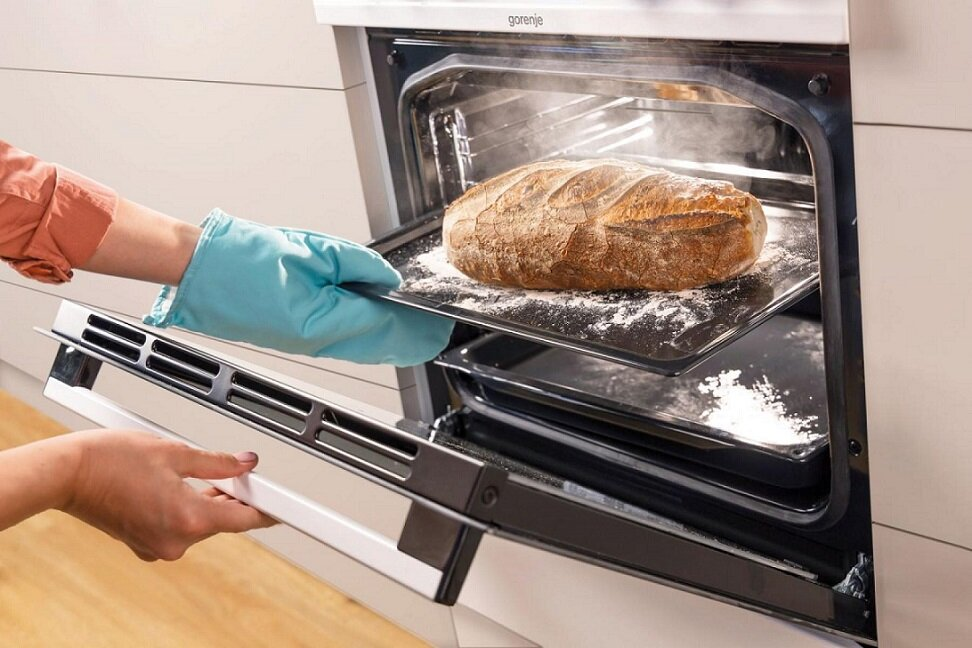 Osoba w rękawicach kuchennych wyjmuje tacę z bochenkiem chleba z otwartego piekarnika.
