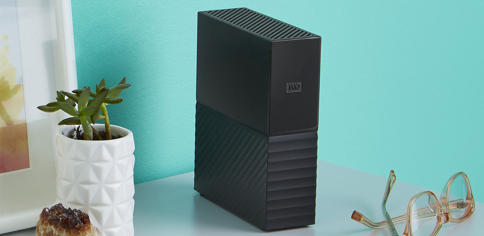 Disque dur externe noir à côté d'un pot blanc, d'une petite plante succulente et de lunettes.