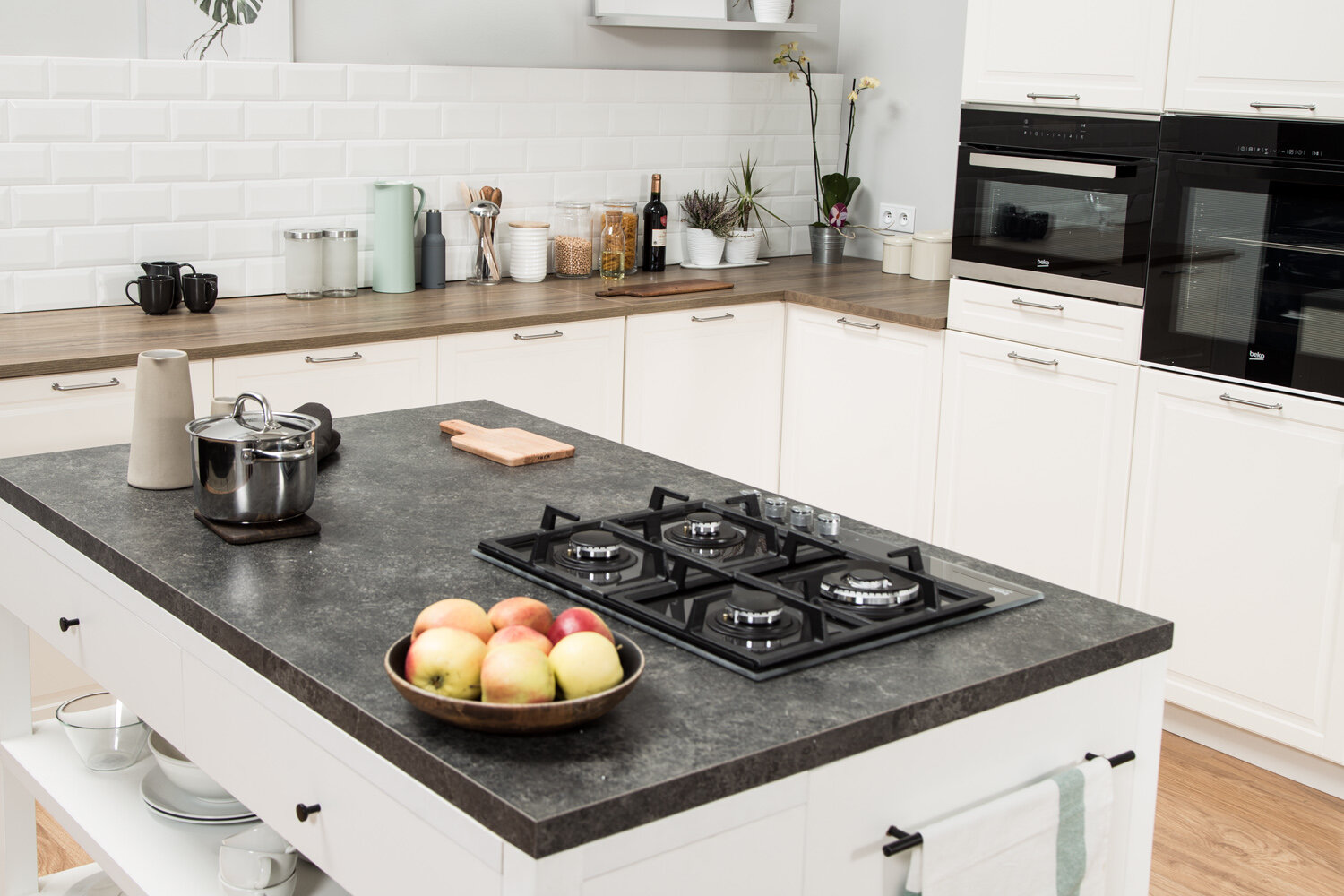 Wnętrze kuchni z wyspą z płytą grzewczą i miską jabłek. Białe szafki i biała ceglana ściana.