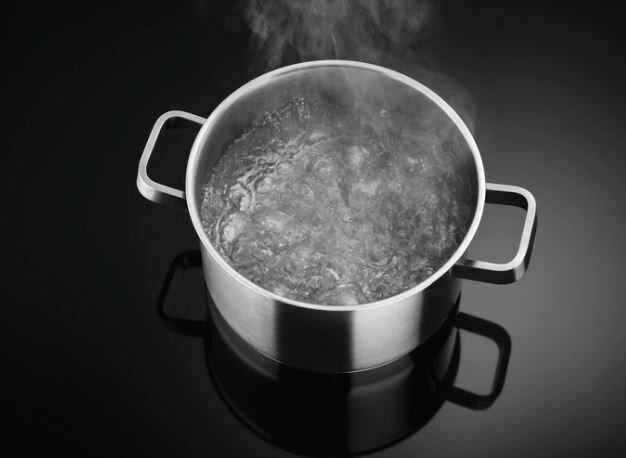 Garnek ze stali nierdzewnej z wrzącą wodą na czarnej płycie grzewczej, para unosi się nad nim.
