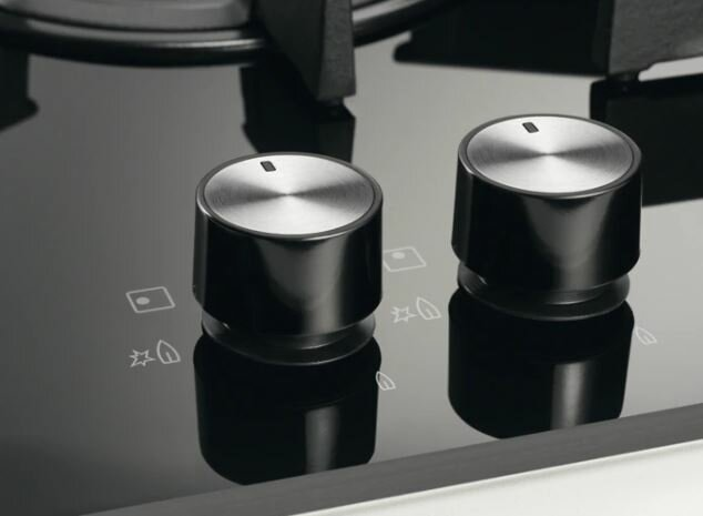 Zbliżenie czarnych pokręteł kuchenki z metalowymi nakładkami na czarnej powierzchni.