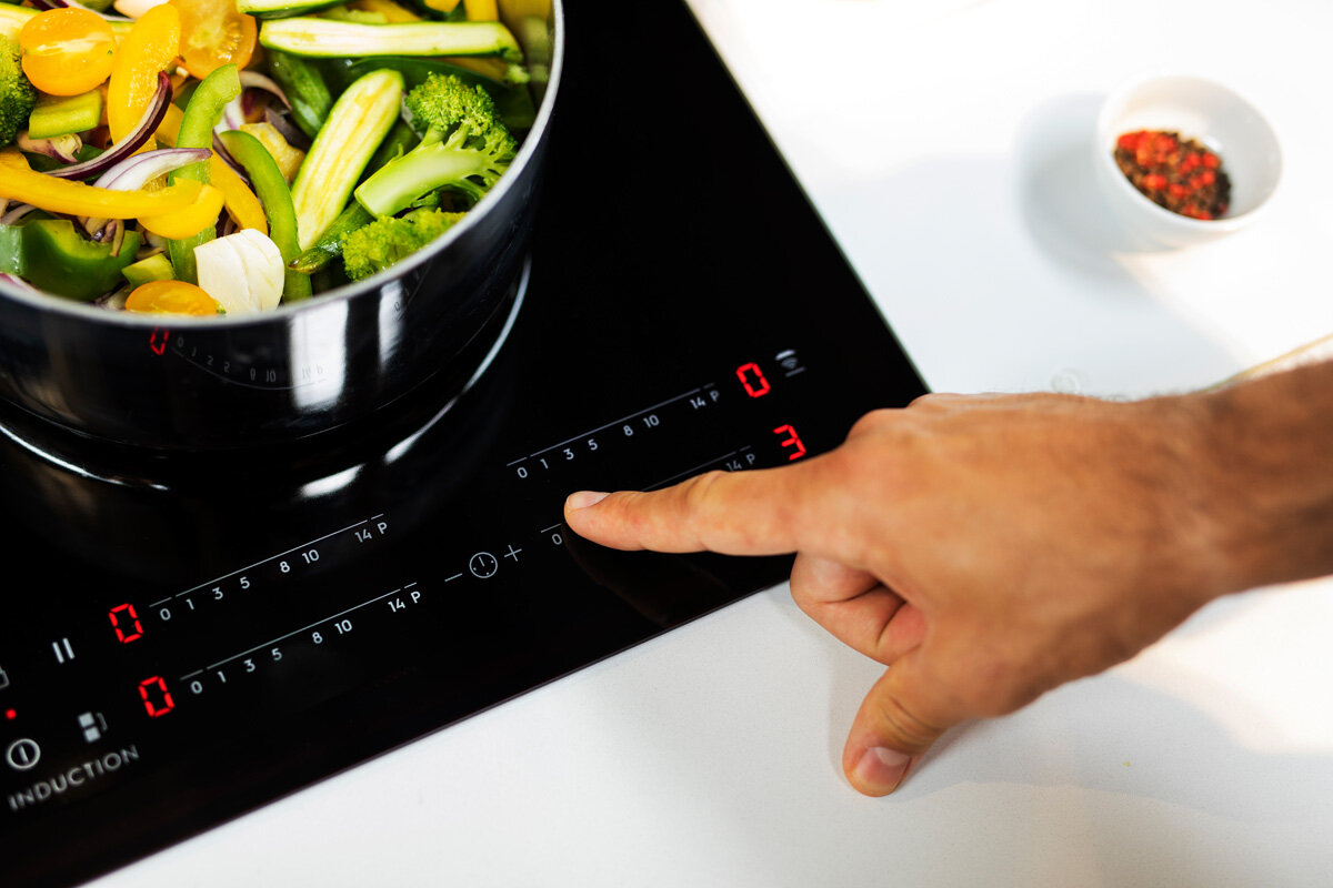 Ręka naciskająca przycisk na czarnej płycie indukcyjnej z garnkiem z jedzeniem.