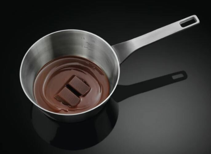 Garnek ze stali nierdzewnej z roztopioną czekoladą i dwoma kwadratami czekolady w środku, na czarnej powierzchni.