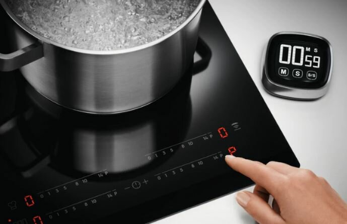 Czarna płyta grzewcza z garnkiem wrzącej wody i timerem pokazującym 00:59. Palec dotyka przycisku.