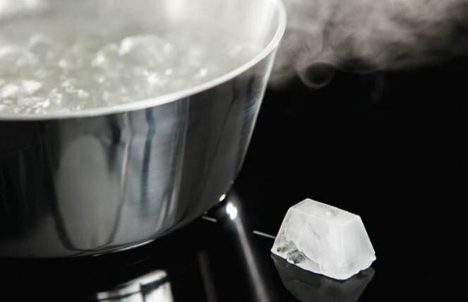 Garnek ze stali nierdzewnej z wrzącą wodą. Kostka lodu leży na czarnej powierzchni.