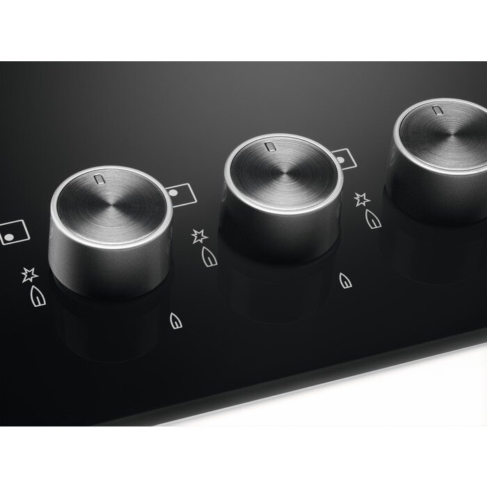 Zbliżenie trzech srebrnych pokręteł na czarnej płycie kuchennej. Widoczne symbole płomieni i palników.