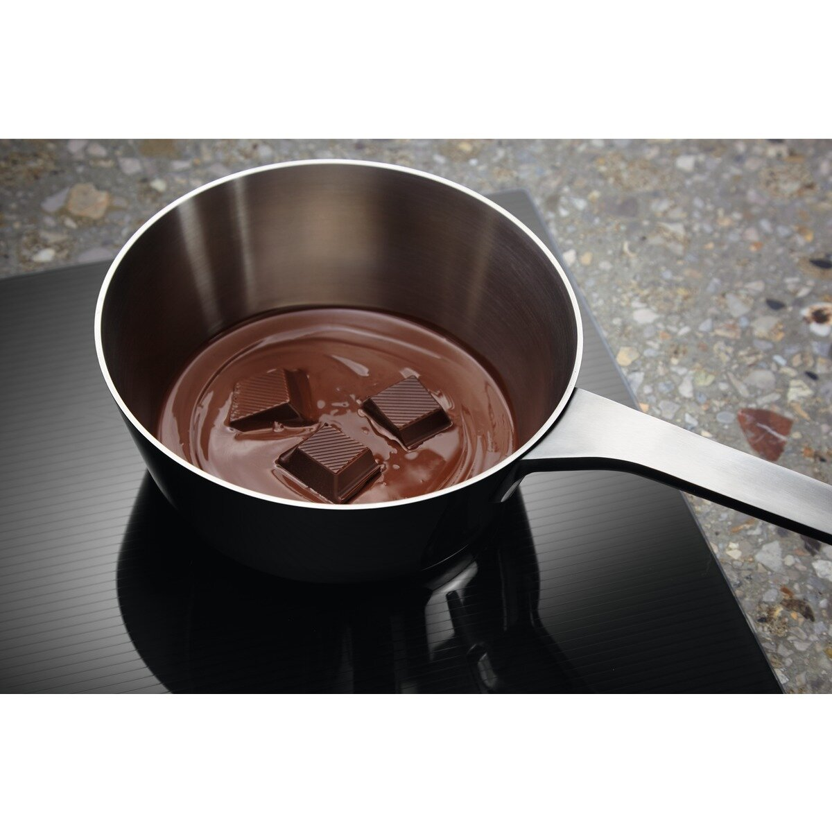 Garnek z roztopioną czekoladą i trzema kwadratami czekolady na czarnej płycie indukcyjnej.