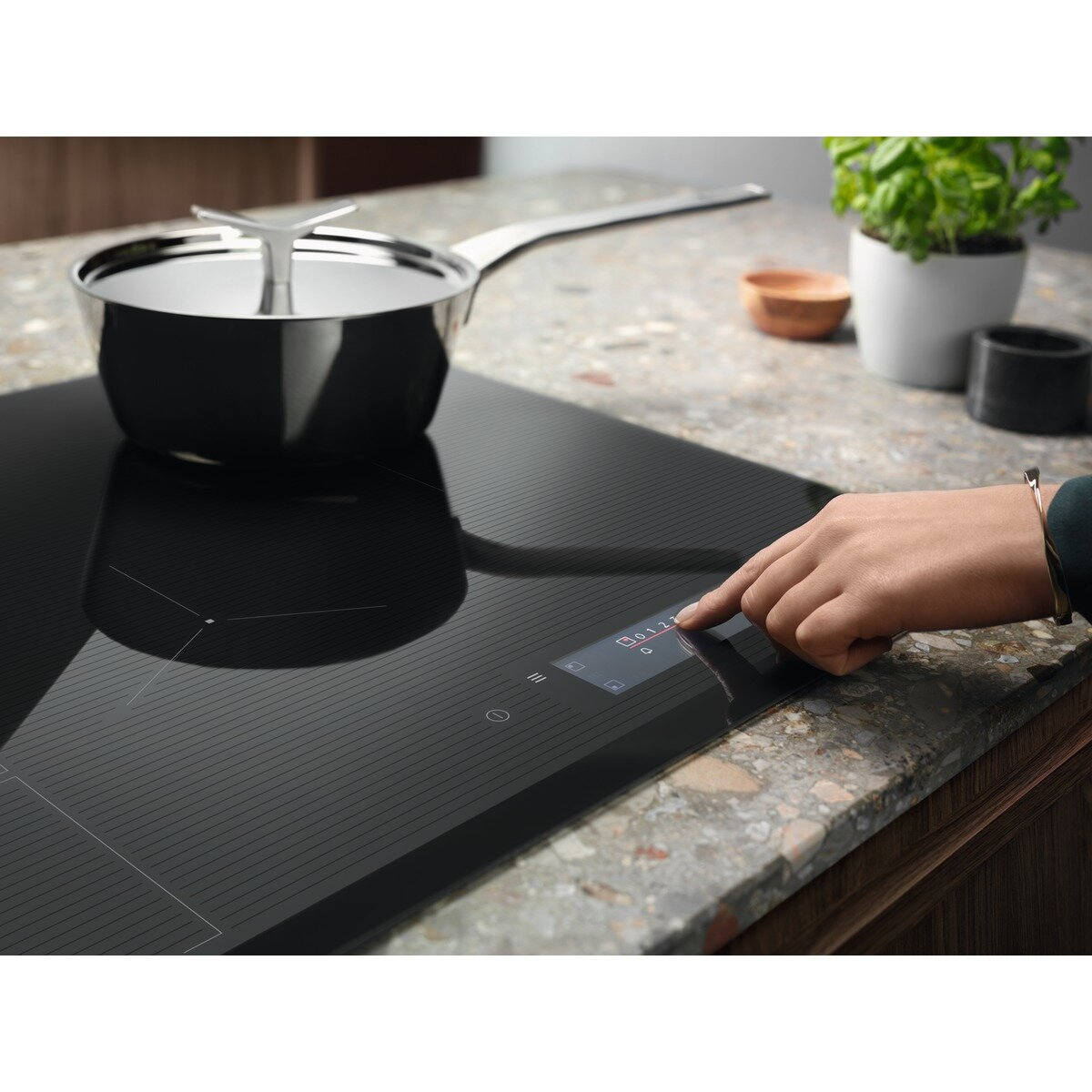 Dłoń dotykająca cyfrowego wyświetlacza na czarnej płycie indukcyjnej z garnkiem.