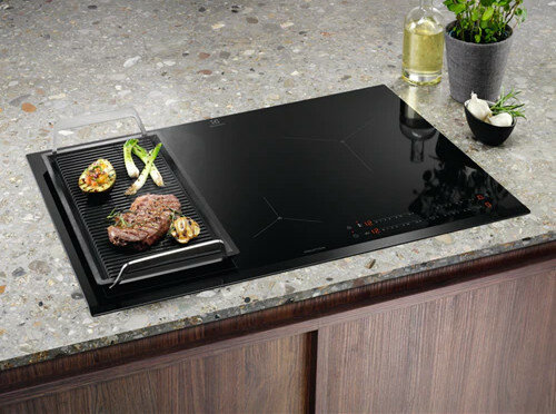 Czarna płyta indukcyjna z grillem i jedzeniem. Jest na blacie.