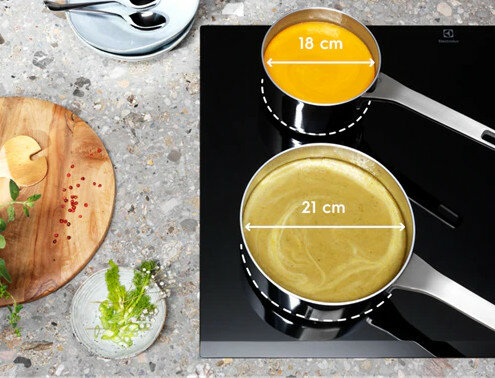 Dwa garnki na czarnej płycie, jeden z pomarańczową zupą, 18cm, drugi z zieloną, 21cm.
