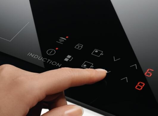 Dłoń dotyka czarnej płyty indukcyjnej. Wyświetlacz pokazuje symbole kontrolne i cyfry cyfrowe w kolorze czerwonym.