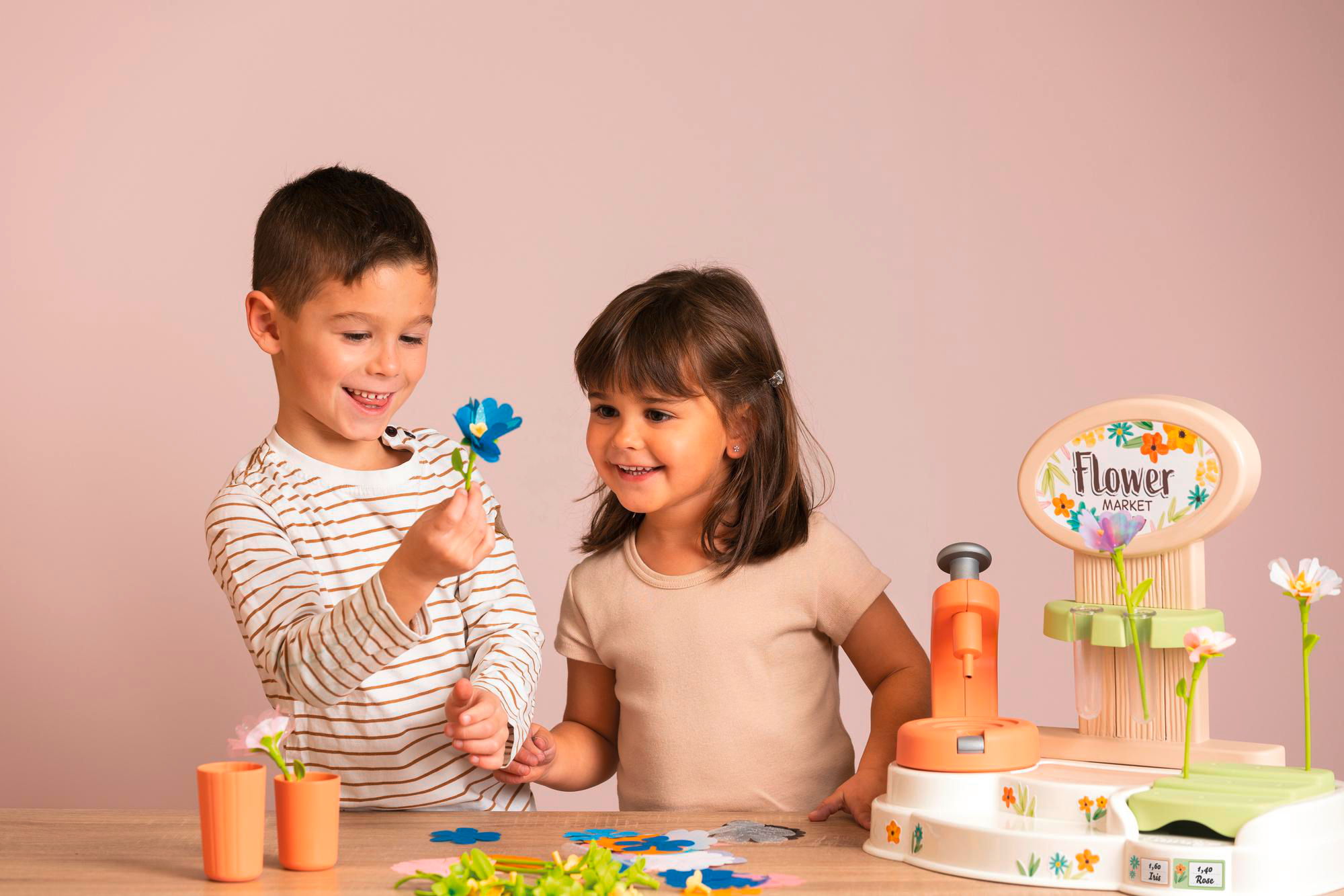 Zwei Kinder spielen mit einem Spielzeug-Blumenmarkt. Junge hält blaue Blume, Mädchen lächelt. Rosa Hintergrund.