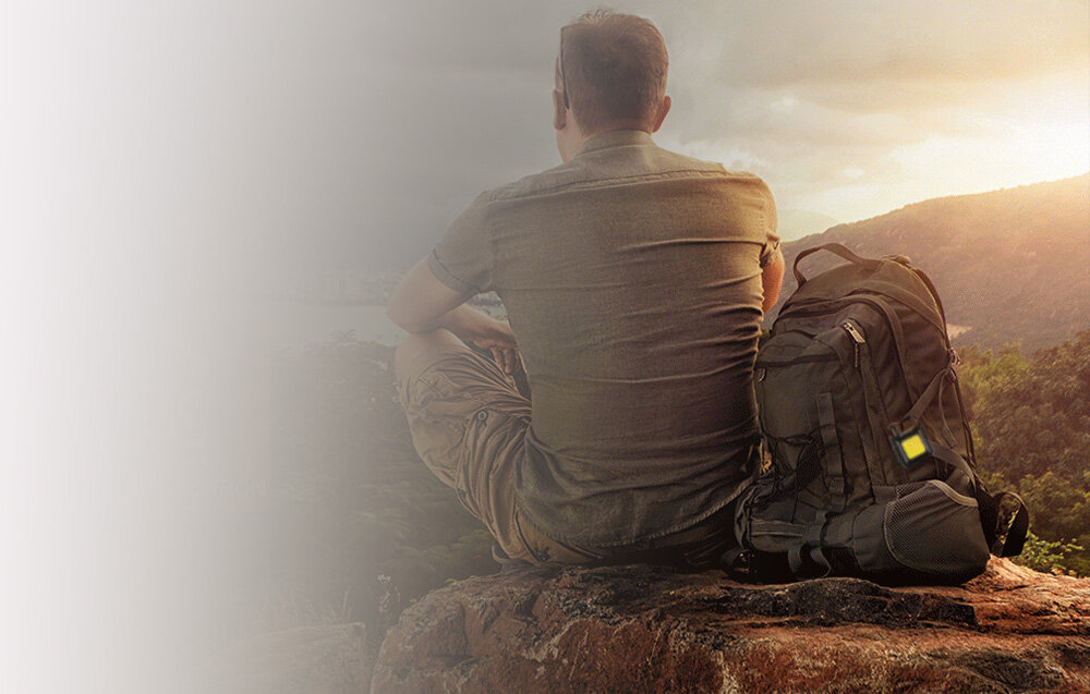 Mężczyzna siedzi na skale i patrzy na pasmo gór podczas zachodu słońca. Obok niego plecak.
