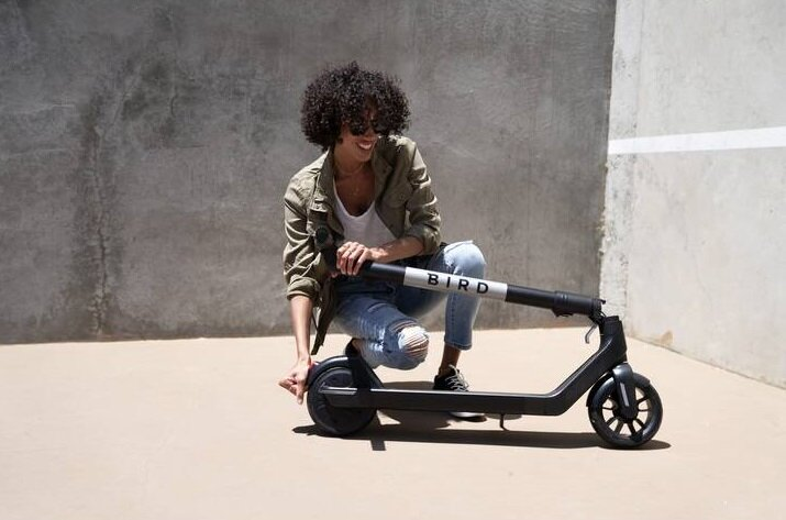 Kobieta z kręconymi włosami, uśmiechnięta, klęcząca przy czarnej hulajnodze Bird.