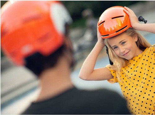 Blondynka zakłada pomarańczowy kask. Nosi żółtą sukienkę w kropki.