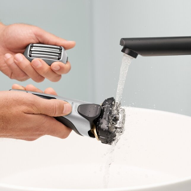 Mężczyzna myje golarkę pod kranem. Golarka jest srebrna i czarna. Tłem jest biały zlew.