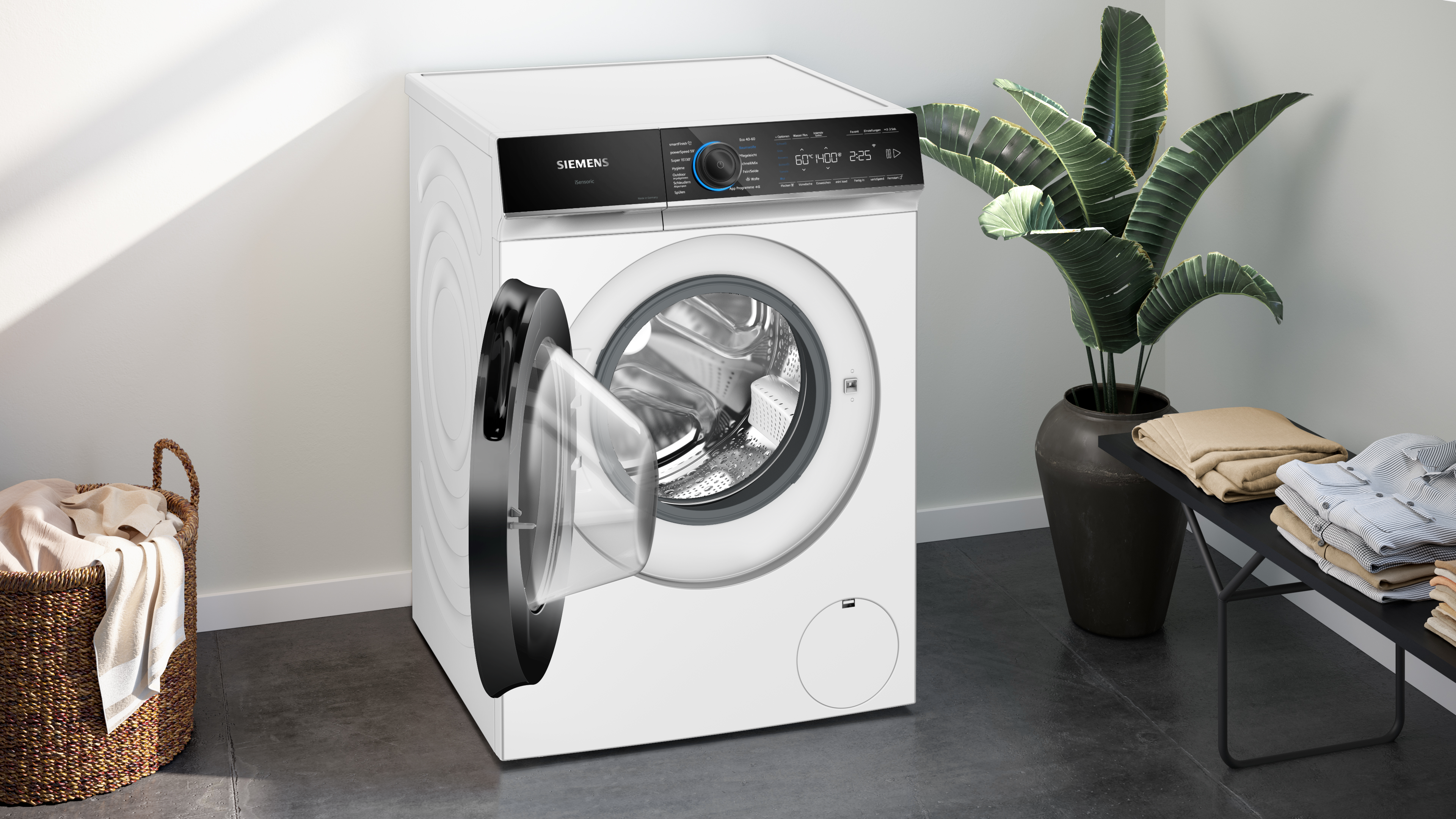 Eine weiße Waschmaschine mit offener Tür, neben einer Pflanze und gefalteter Kleidung.