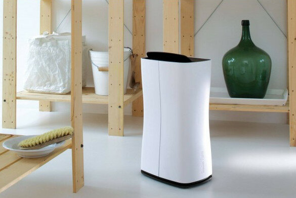Biały oczyszczacz powietrza z czarnym blatem. Drewniane półki i zielona butelka w tle.