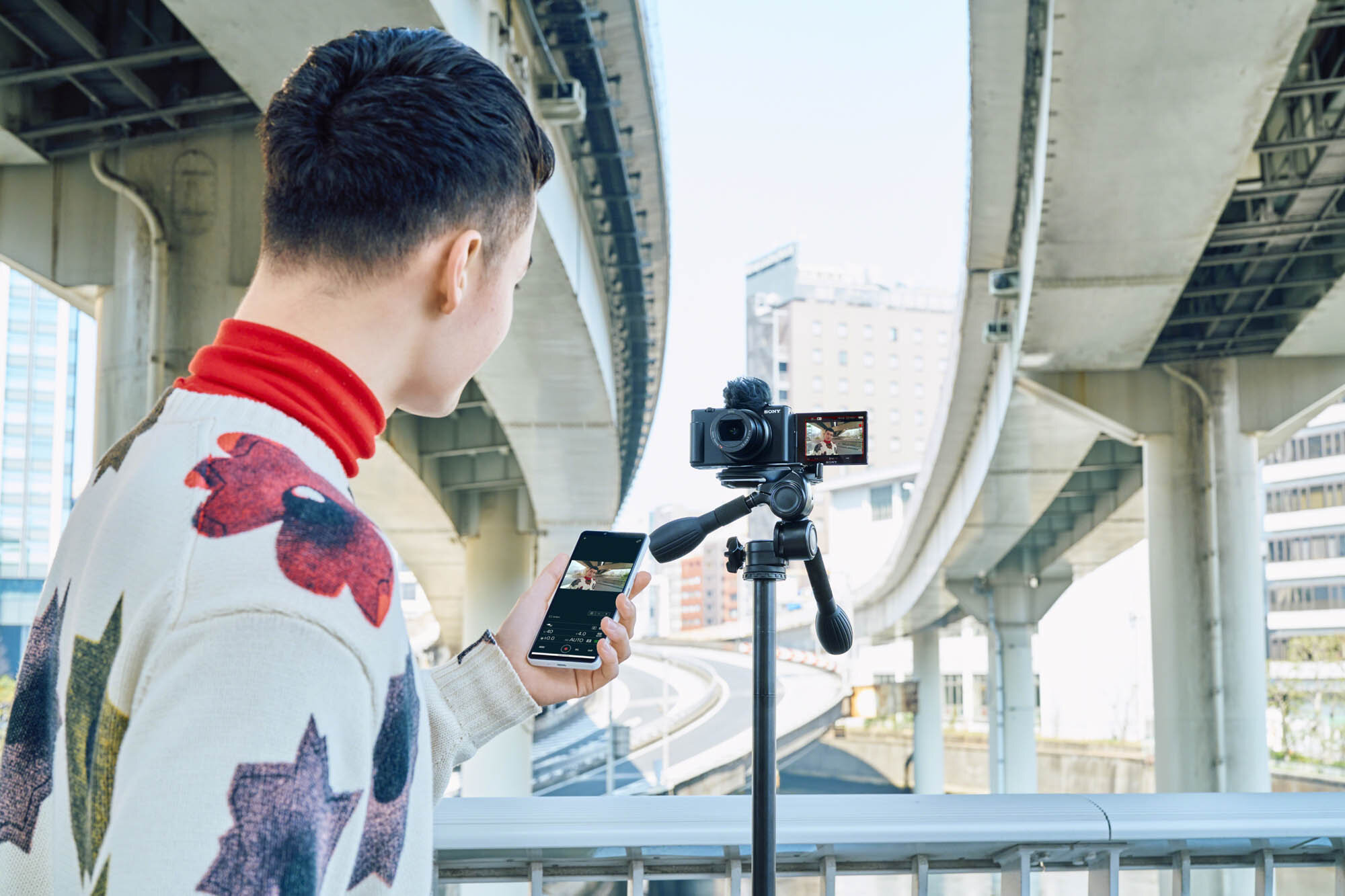 Mężczyzna filmujący kamerą na statywie, trzymający telefon pod mostem.