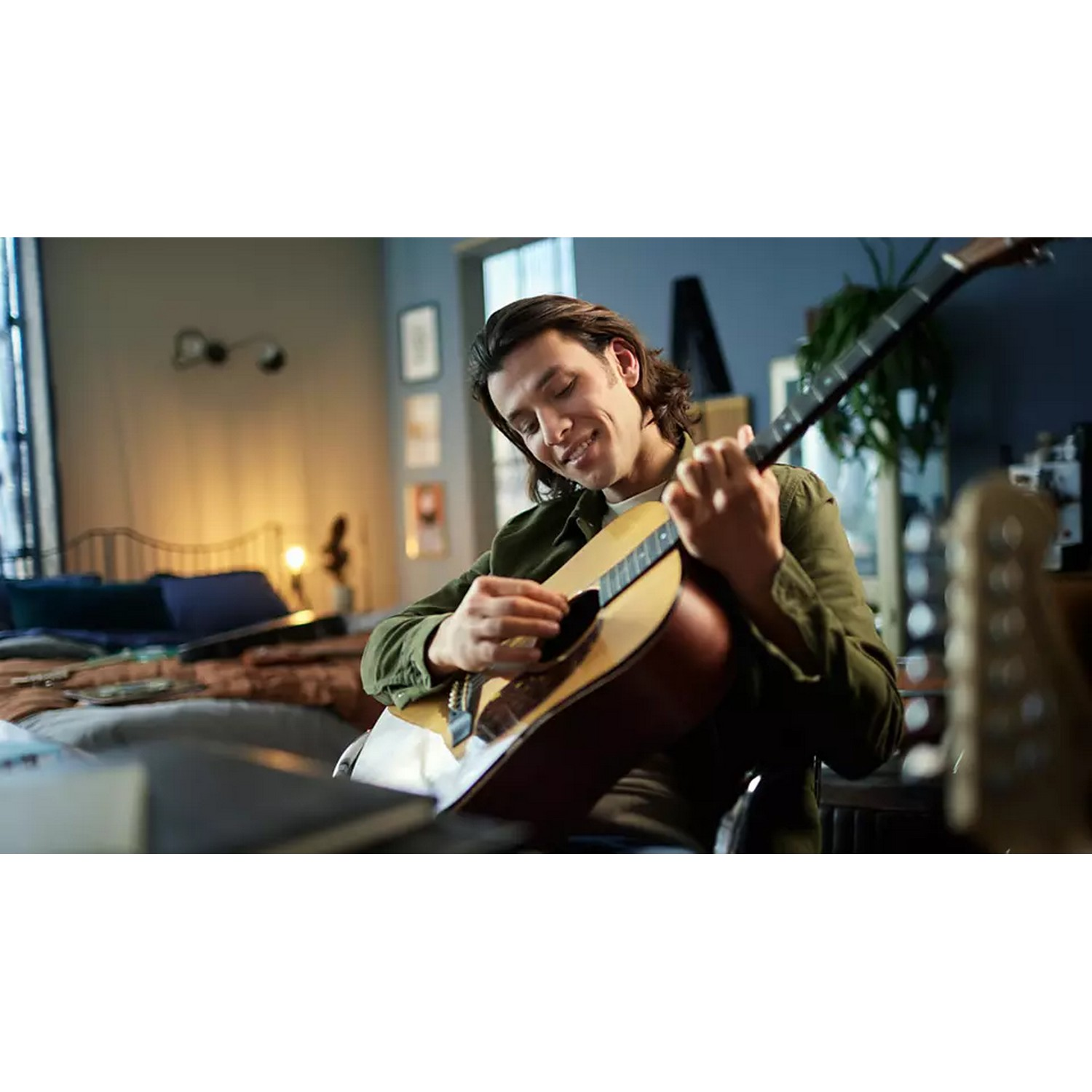 Man plays an acoustic guitar, smiling, with a bedroom scene in the background.