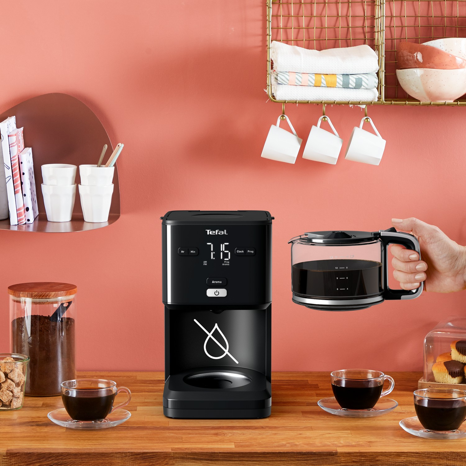 Zwart koffiezetapparaat met een glazen kan en twee kopjes koffie. Roze muur en houten tafel.