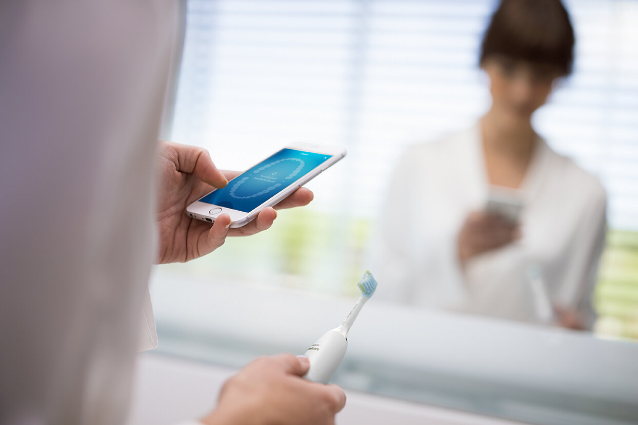 Osoba trzymająca smartfon z aplikacją dentystyczną, szczoteczka do zębów w dłoni. Lustro z rozmytą osobą.