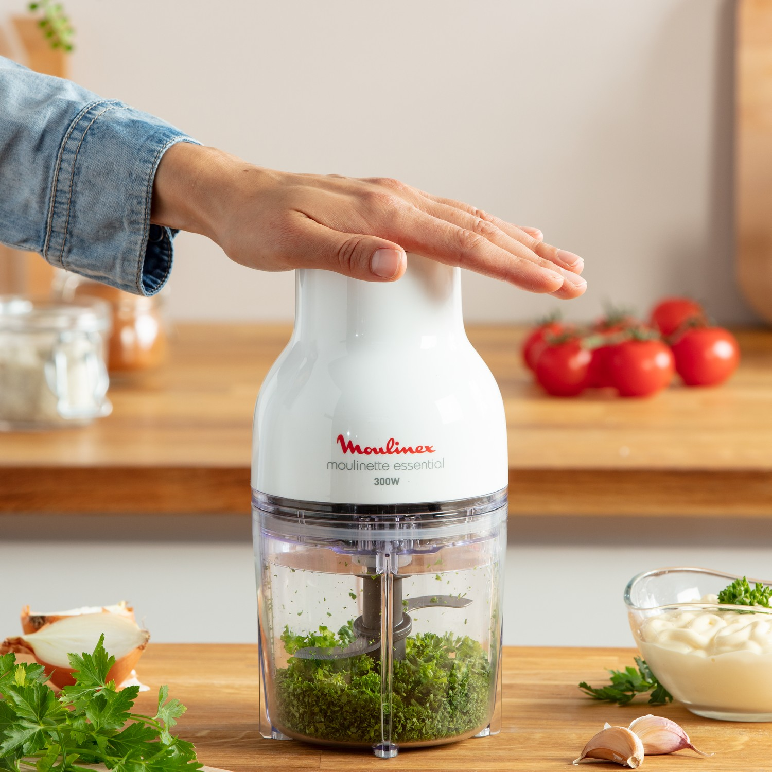 La main d'une personne au-dessus d'un hachoir Moulinex blanc. Des légumes et des ingrédients sont visibles.