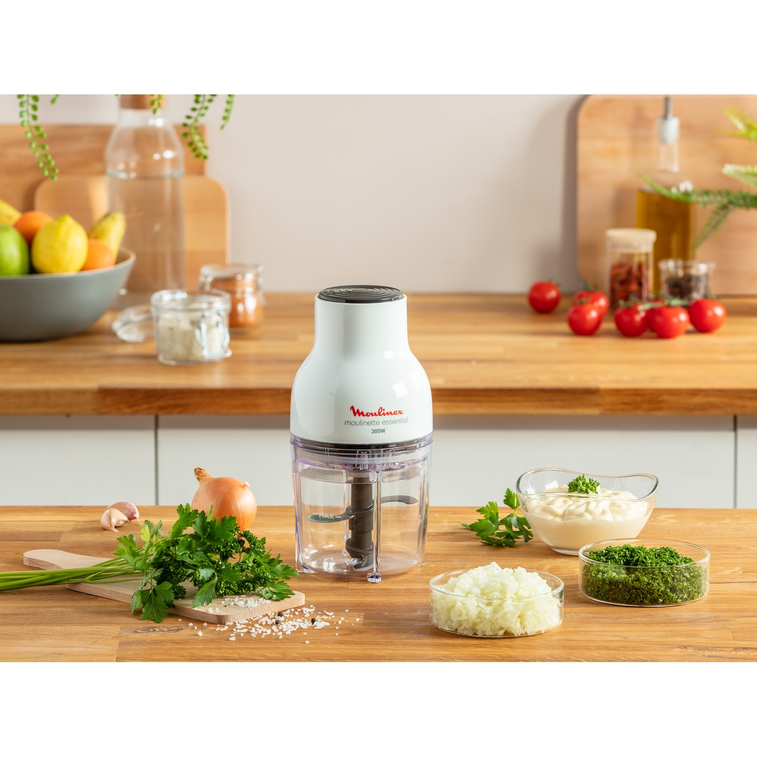Hachoir alimentaire Moulinex blanc avec ingrédients sur une table en bois. Inclut persil, oignon et tomates.