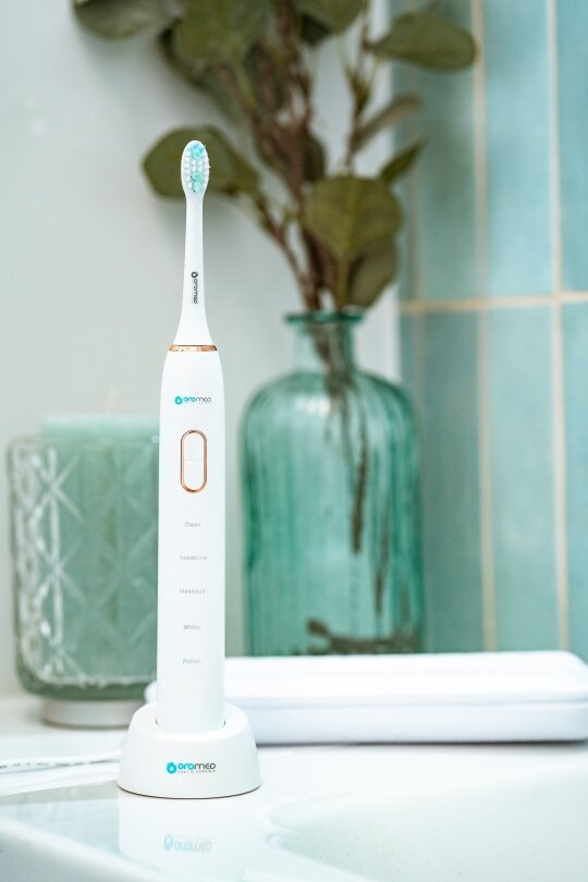 Biała elektryczna szczoteczka do zębów na podstawce. Za nią wazon z roślinami.