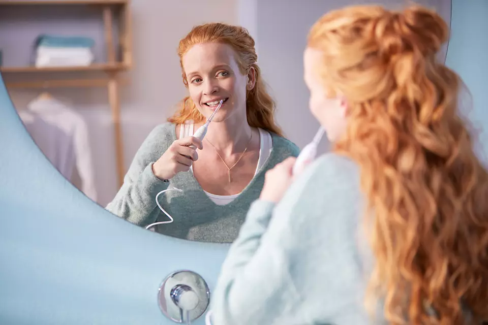 Kobieta używa irygatora dentystycznego przed lustrem. Ma na sobie zieloną górę.