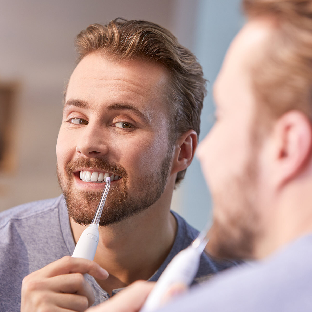 Mężczyzna uśmiecha się do lustra, używając szczoteczki Philips Sonicare. Odbicie pokazuje urządzenie z bliska.