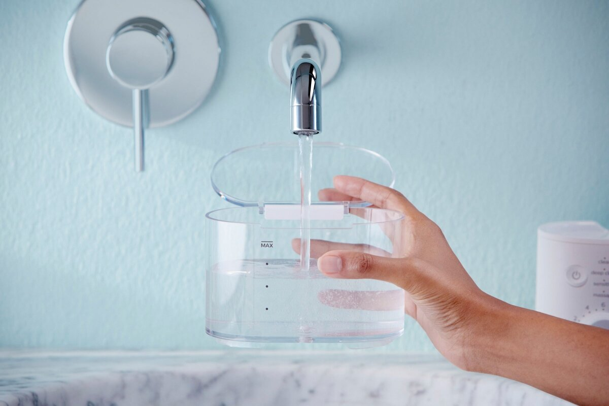 Ręka napełniająca przezroczysty pojemnik na wodę pod kranem. Niebieska ściana w tle.