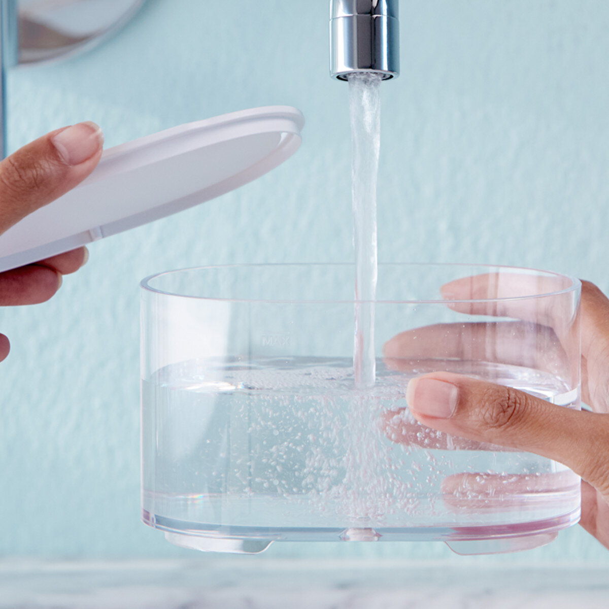 Osoba napełnia przezroczysty pojemnik wodą z kranu. Biała pokrywka znajduje się po lewej stronie pojemnika.