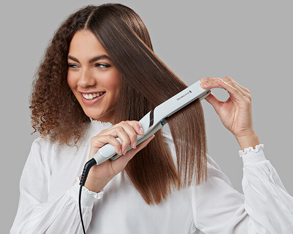 Kobieta używająca białej prostownicy do włosów. Jej włosy są proste z jednej strony, a kręcone z drugiej.