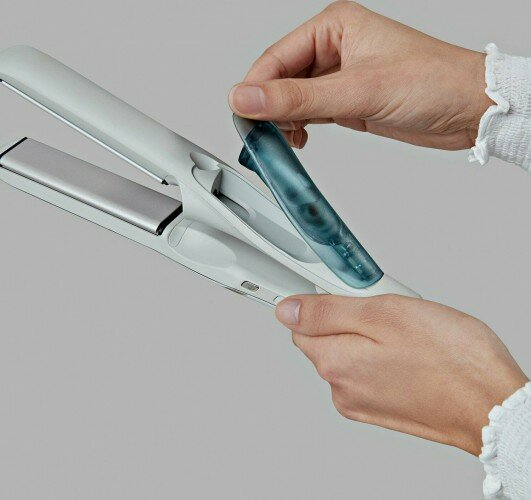 Dłonie używające biało-srebrnej prostownicy do włosów z niebieskim zbiornikiem na wodę.