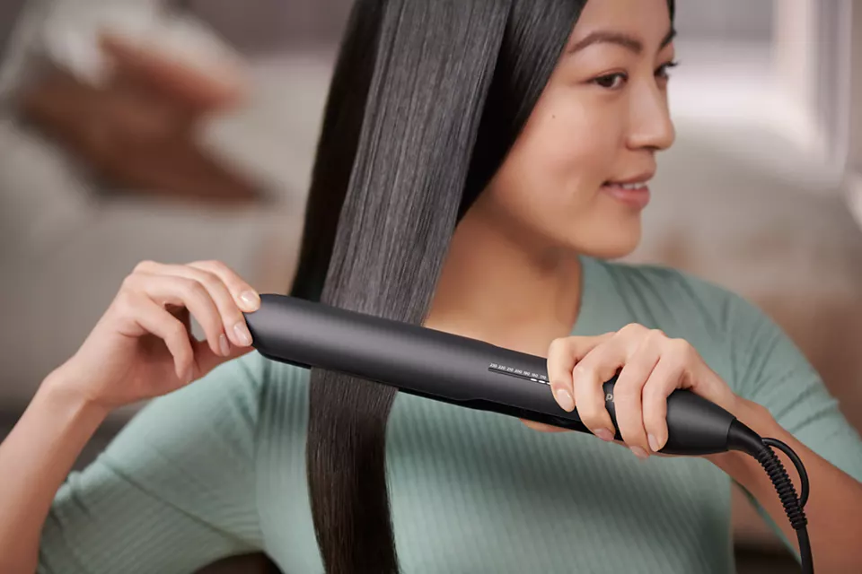 Kobieta prostuje czarną prostownicą ciemne włosy, uśmiechając się. Tło jest rozmyte.