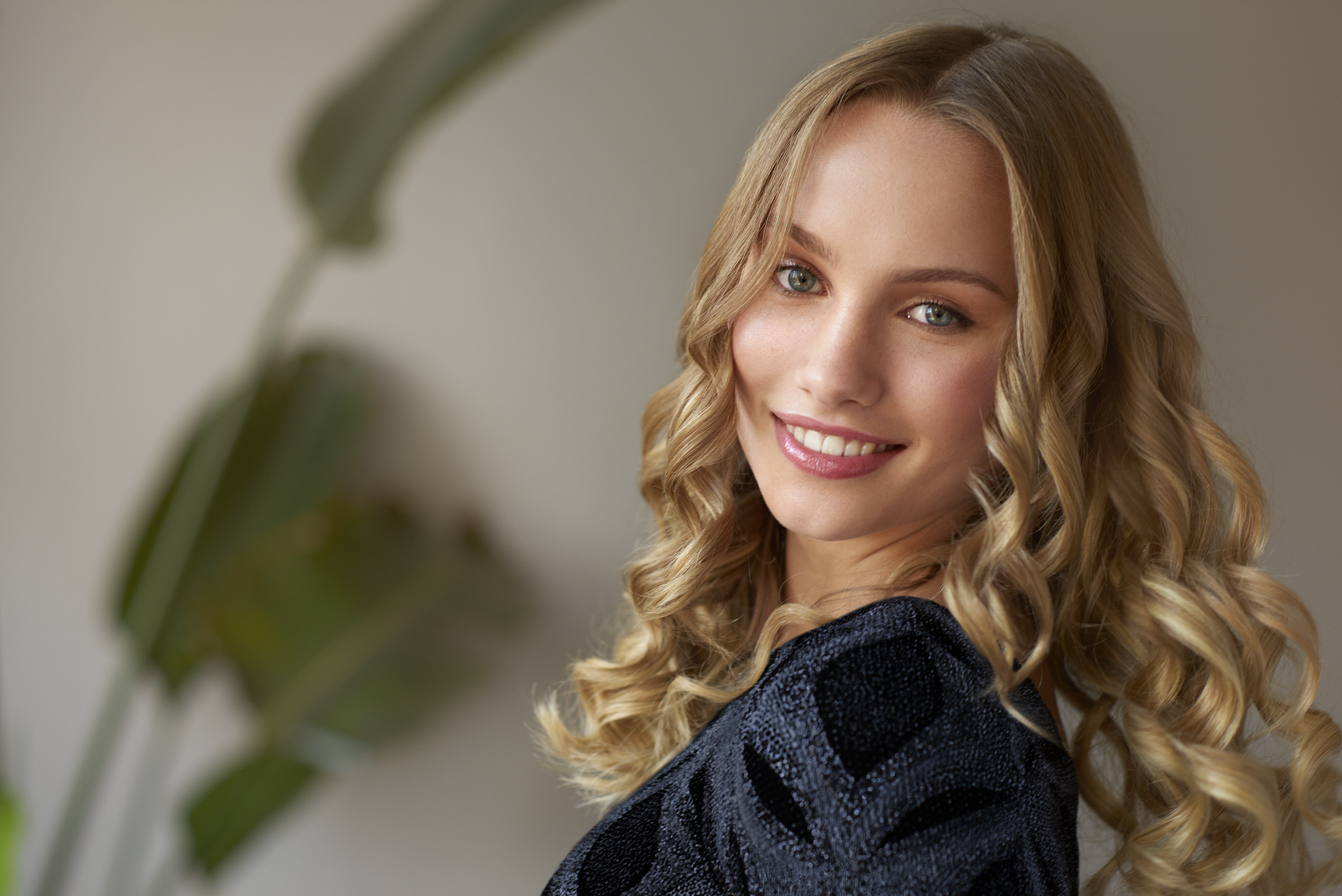 Blondynka uśmiecha się, kręcone włosy, czarny top. Zielona roślina w tle.