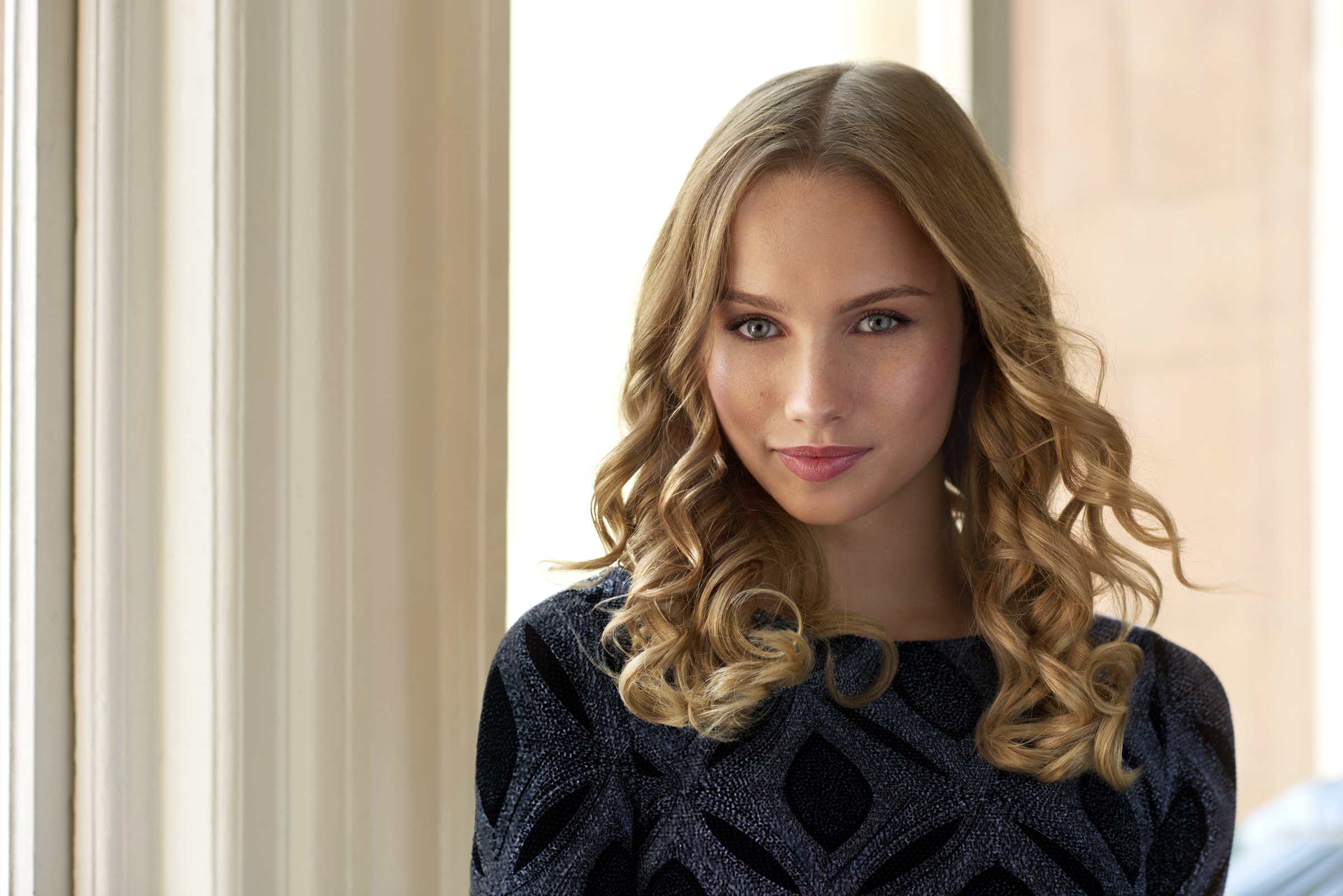 Blondynka z kręconymi włosami, w czarnym topie. Jasne tło, blisko okna.