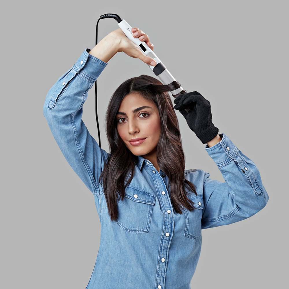 Kobieta kręci włosy lokówką. Ma na sobie dżinsową koszulę i czarną rękawicę. Tło jest szare.