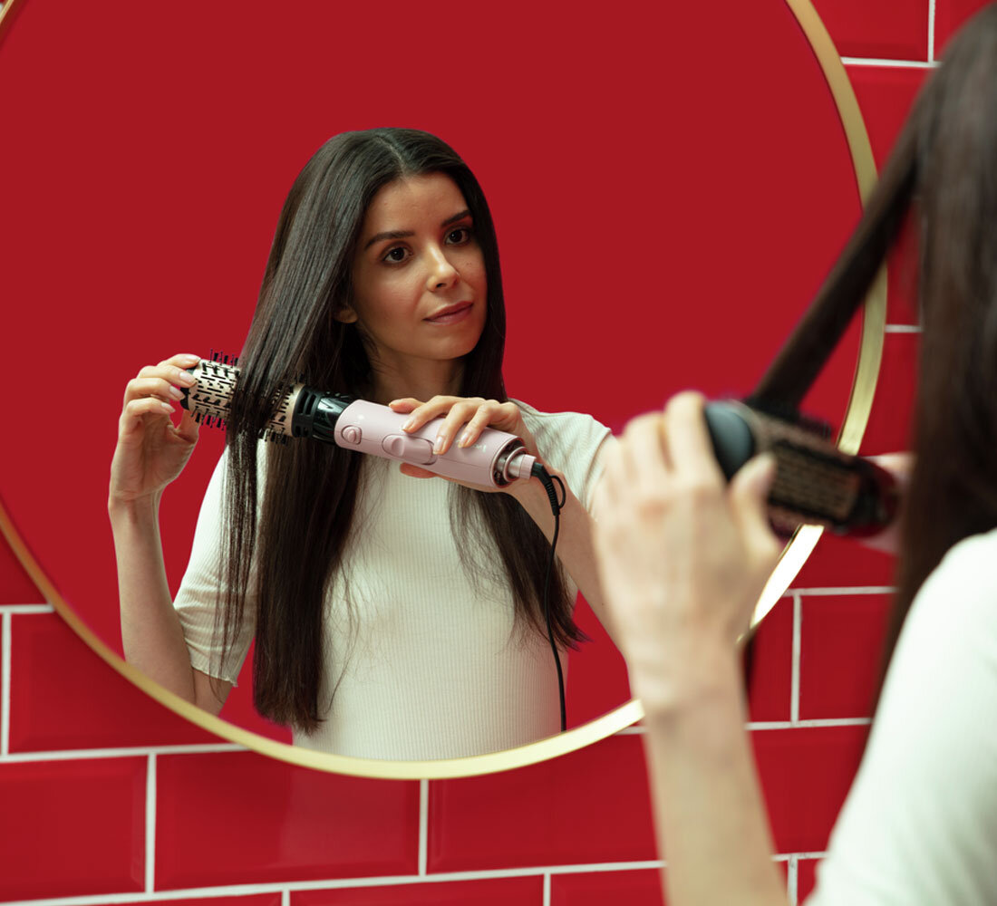 Kobieta stylizuje włosy w lustrze. Używa szczotki po lewej i prostownicy po prawej. Czerwone kafelki w tle.