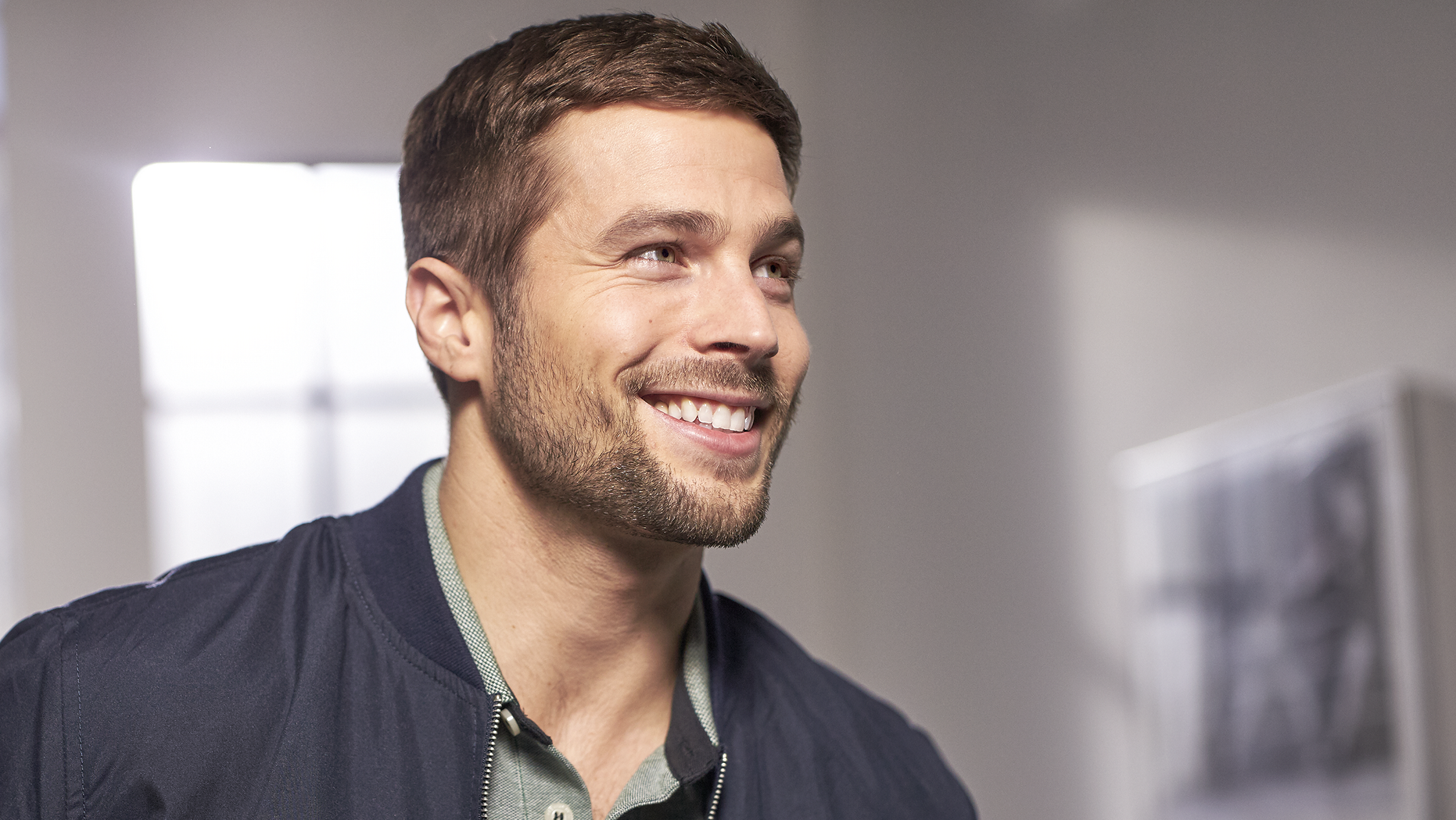 Uśmiechnięty mężczyzna w niebieskiej kurtce. Ma brodę i krótkie brązowe włosy. Jasne tło.