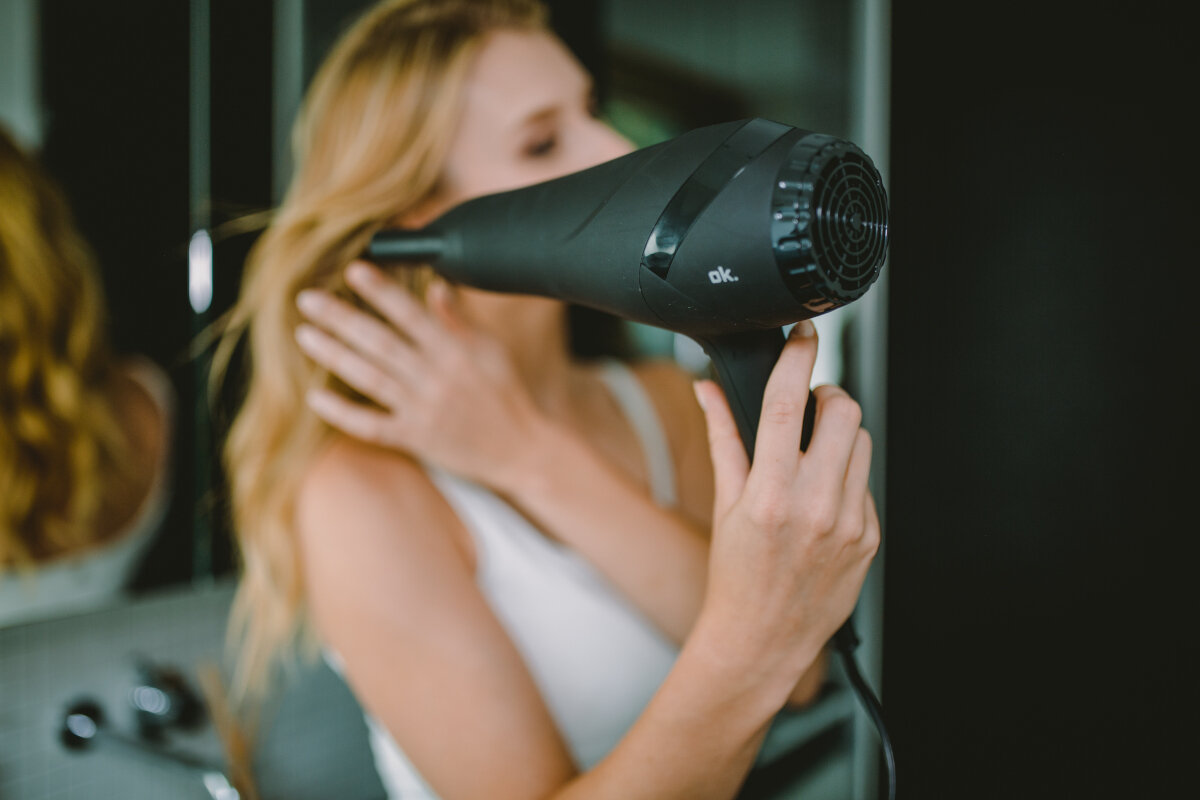 Kobieta używająca czarnej suszarki do włosów na swoich blond włosach, przed lustrem.