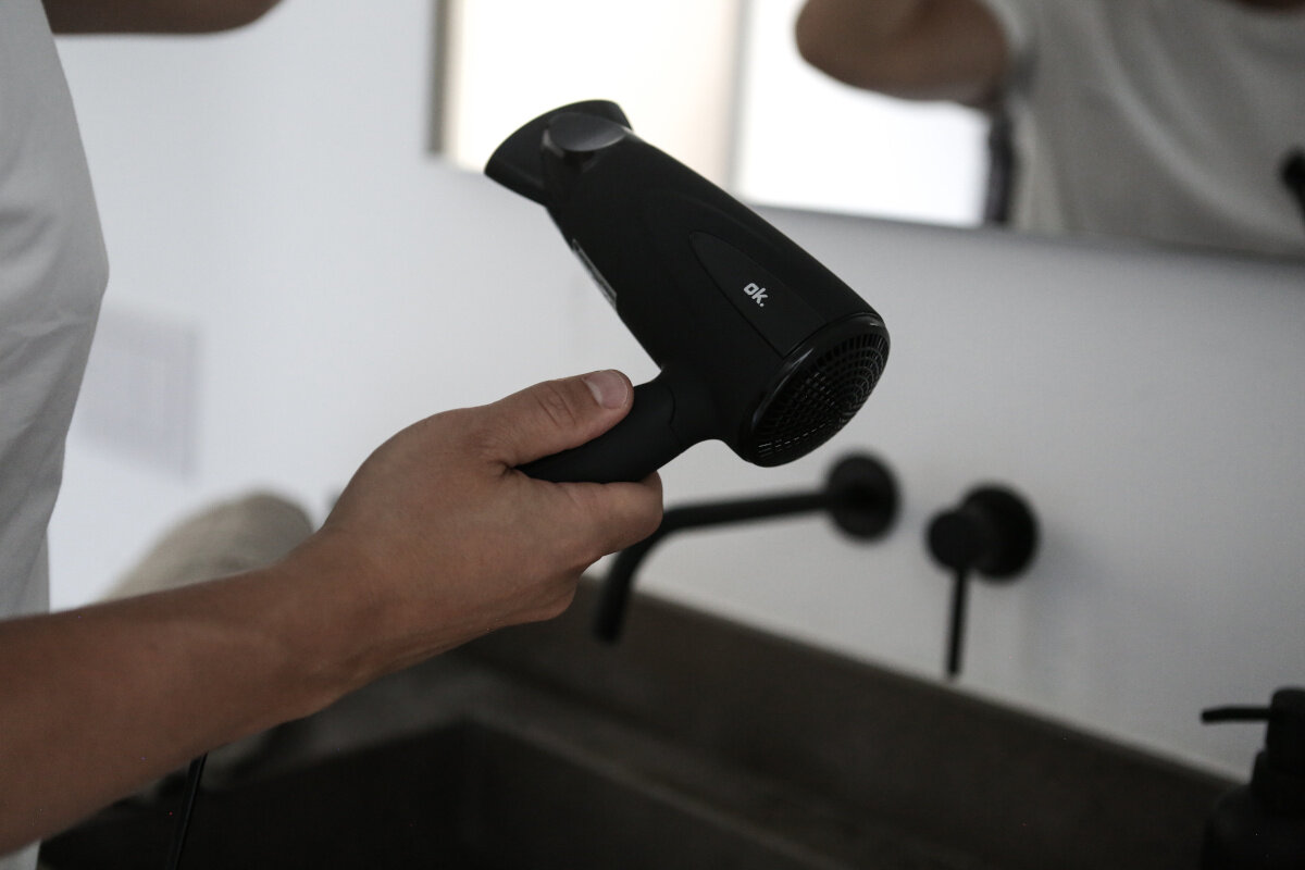 Osoba trzymająca czarną suszarkę przed lustrem i umywalką.