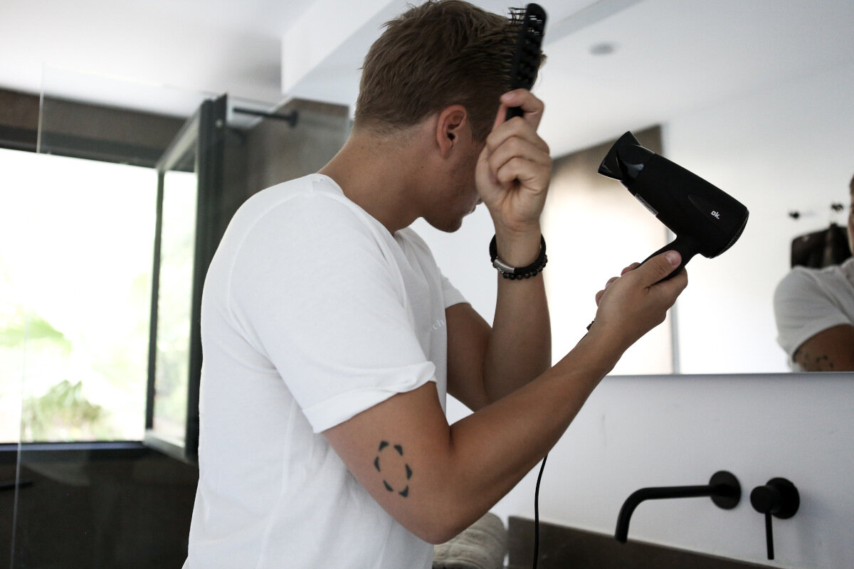 Mężczyzna w białym t-shircie czesze włosy szczotką i trzyma suszarkę do włosów przed lustrem.