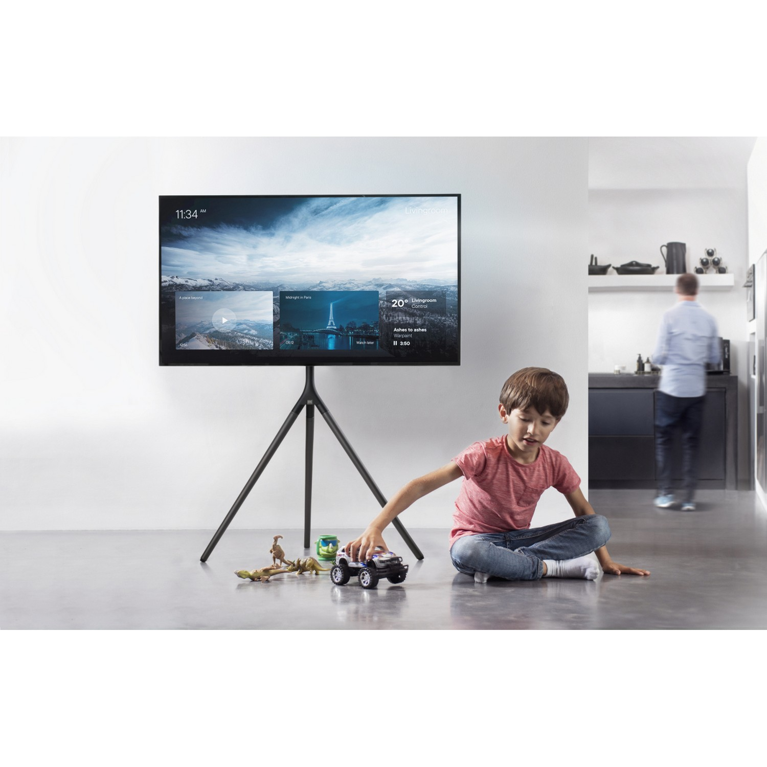 Een kind speelt met speelgoedauto's voor een grote tv. Een man op de achtergrond.