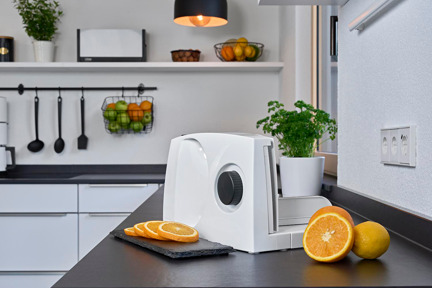 Eine Küchenszene mit einem weißen Lebensmittelschneider. Auf einer schwarzen Oberfläche liegen Orangenscheiben.