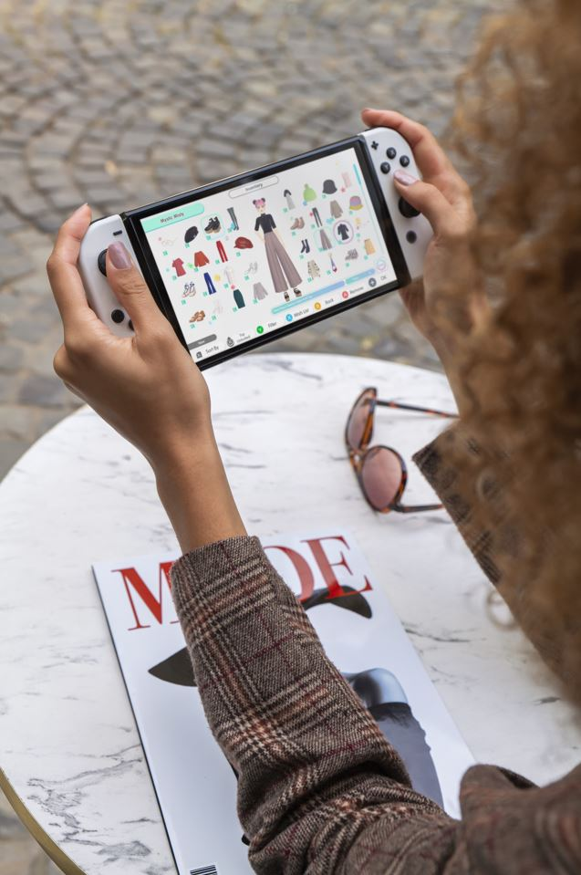 Persoon houdt een witte Nintendo Switch vast, met een garderobe-interface. Tijdschrift en zonnebril op tafel.
